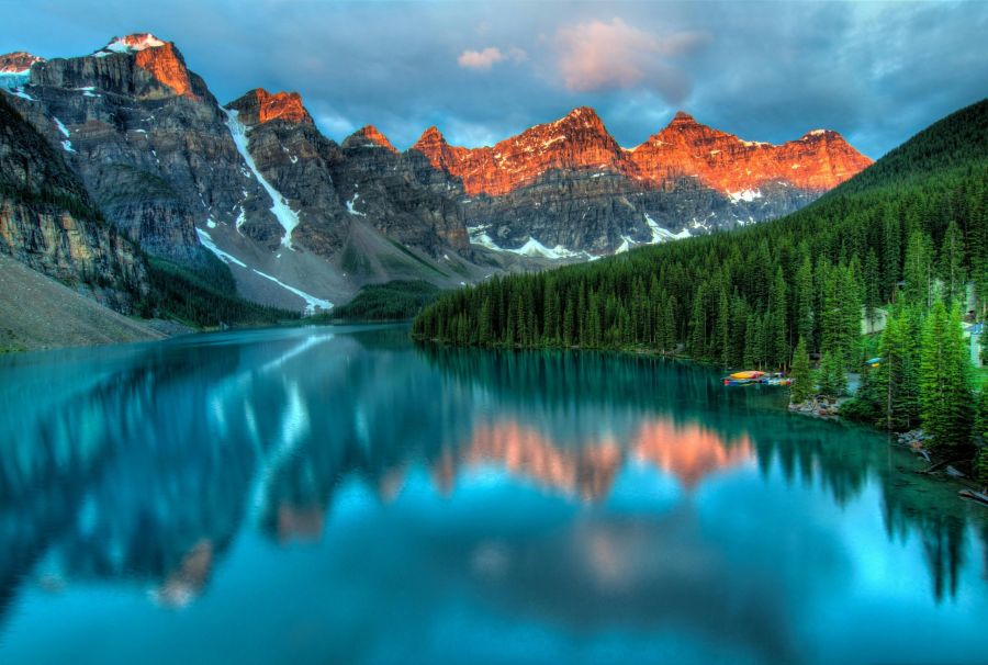A tranquil mountain lake at sunrise, reflecting snow-capped peaks and evergreen trees, with golden light casting a warm glow on the rugged cliffs in the background.
